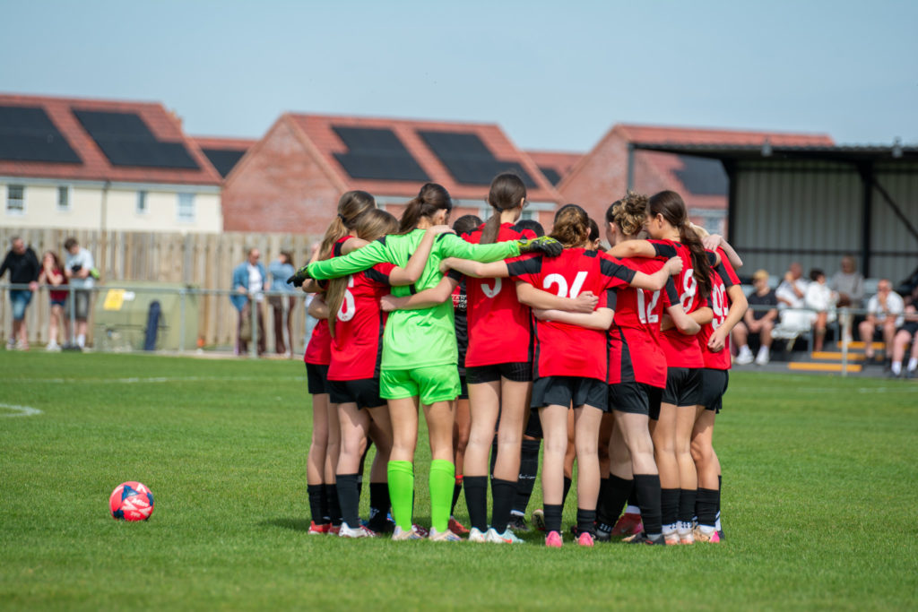 Group of girls on a football pitch in a huddle playing a football game.